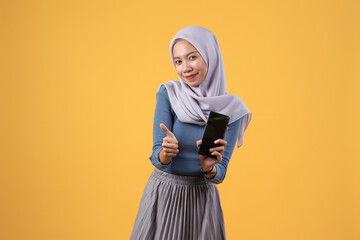 happy asian indonesian muslim woman holding smart phone showing blank screen and giving thumb up gesture on isolated yellow background