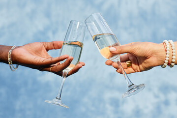 Two hands belonging to diverse individuals clinking champagne glasses against blue backdrop,...