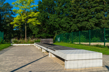 A sleek concrete bench sits in a peaceful park, bathed in sunlight with vibrant green trees in the background, inviting visitors to relax and enjoy nature.