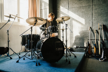 Fototapeta premium Drummer perfecting his skills in brightly sunlit music studio, he plays drum set. Various guitars rest against wall.