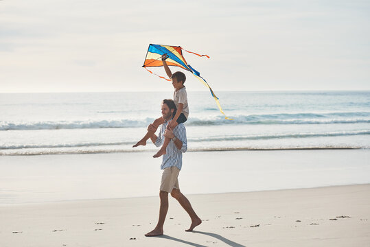 Father, boy and walking on beach with kite, water and carrying for travel, holiday and bonding as fun. Man, son and shoulder ride by ocean with support, love and care for trip, vacation and playing