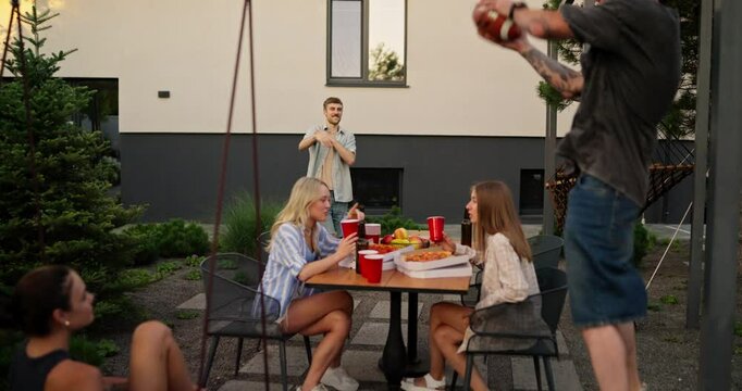 Happy blond guy with a mustache throws an American football ball to his friend while relaxing at a table in the backyard of a house