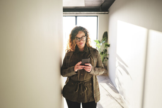 Smiling businesswoman reading a text message in an office hallway. She wears black glasses and has big curly hair. The sun comes in through a window behind her.