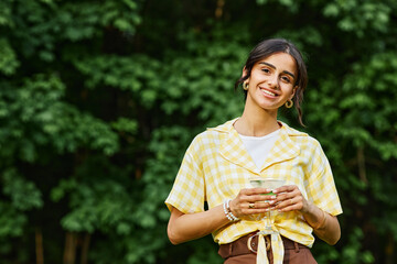 Smiling young woman enjoying outdoor setting with lush greenery in background, wearing checkered shirt and holding beverage