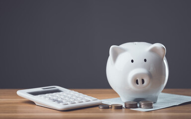White piggy bank, bank book, and a pile of coins rest on a wooden table against a dark gray background. The scene symbolizes financial planning, savings, and the importance of managing money wisely.