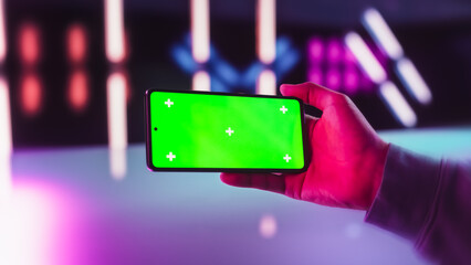 Gamer Person Checking a Social Network Post on Smartphone with Mock Up Green Screen Chromakey Display. Person Holding Cellphone Horizontally in a Futuristic Neon Room, Streaming Entertainment Content © Gorodenkoff
