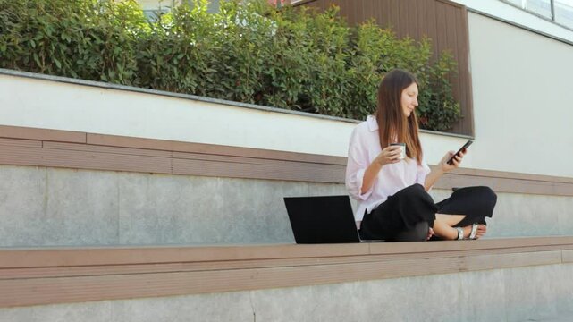 Woman working on laptop in park. Cute female freelancer making remote work and drinking tea in open air. 