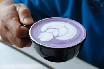 Close-up of a hand holding a cup of purple latte art, featuring a detailed leaf design. Perfect for use in cafe promotions, coffee culture blogs, social media, and lifestyle content.