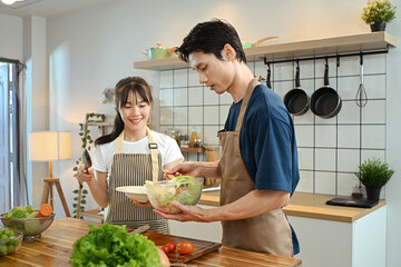 Happy and beautiful young couple wearing aprons preparing healthy vegan salad in the kitchen