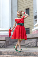 Blonde girl in a red dress on the street with an umbrella near a building on a walk on a summer day