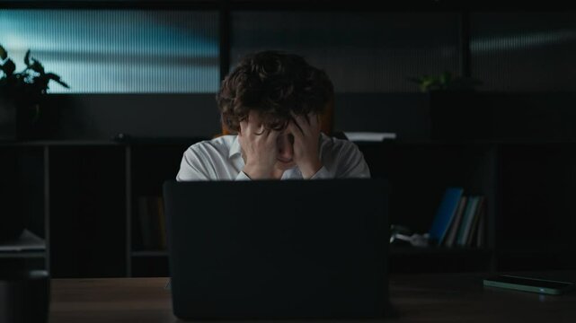 A guy with curly hair in a white shirt covers his face with his hands and shakes his head to the sides during a difficult task at work in the office