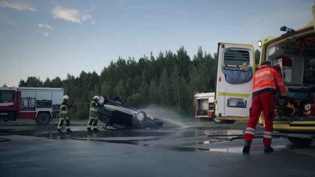 Ambulance Staff Loading Traffic Car Accident Victim into the Vehicle on the Side of the Highway. Firefighters in Protective Uniform with High Pressure Hoses Extinguish the Fire on a Rolled Compact Car
