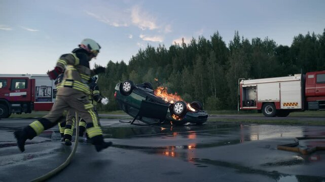 Horrific Traffic Accident with an Upside Down Vehicle: Emergency Services Medical Crew Taking the Patient on a Stretcher to the Ambulance. Firemen with Hoses Start to Extinguish the Fire
