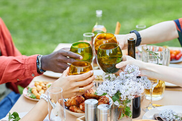 Group of people clinking glasses while enjoying an outdoor meal on a table filled with delicious food and drinks surrounded by fresh-looking greenery