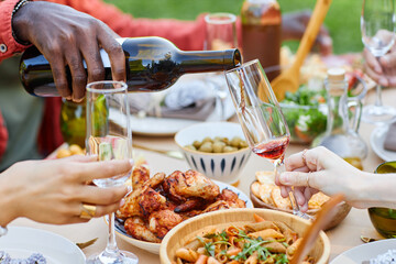 Hands raising wine glasses while dining outdoors with assorted foods including chicken wings, breadsticks, and salads. Image captures convivial atmosphere of shared meal