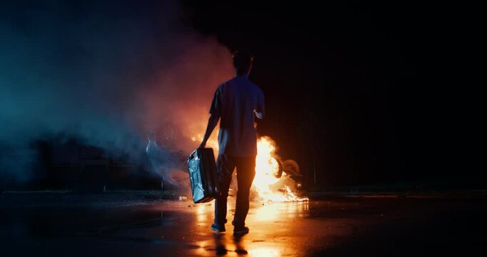 Silhouette of an Anonymous Arsonist Standing with His Back to Camera at Night, Looking at a Burning Vehicle, Holding a Gasoline Canister. Dramatic Scene with a Male Setting a Car on Fire
