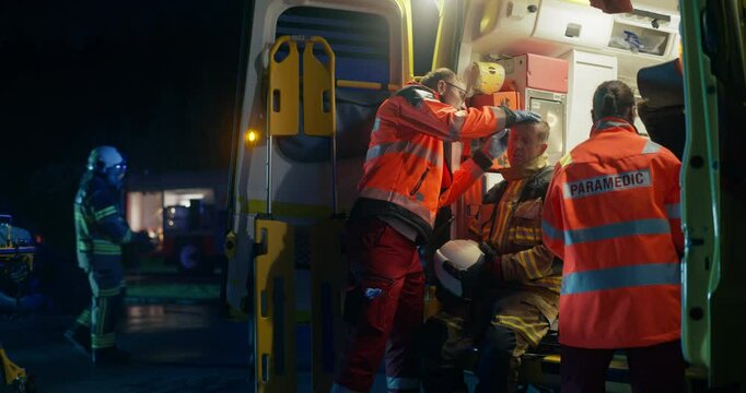Rescue Scene at Night with a Middle Aged Paramedic Diagnosing Injuries on a Male Firefighter with a Flashlight in an Ambulance Vehicle while Firemen Crew Finish Extinguishing a Fire