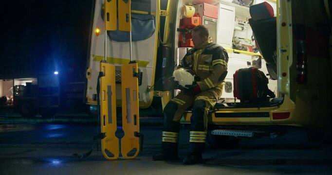 Tired Firefighter Taking Off His Helmet, Sitting Down on the Back of an Ambulance, Resting After a Extinguishing a Hard Fire. Heroic First Responder Relaxing After a Night Traffic Accident Emergency