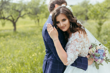 A bride and groom are hugging in a field. The bride is wearing a white dress and the groom is wearing a blue suit. The bride is holding a bouquet of flowers