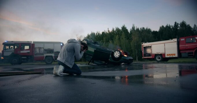 Distressed Young Driver Sitting on the Side of the Road, Crying, Can't Believe He Survived a Horrific Traffic Accident. His Compact Car Lying Upside Down on Asphalt, Bursting in Flames