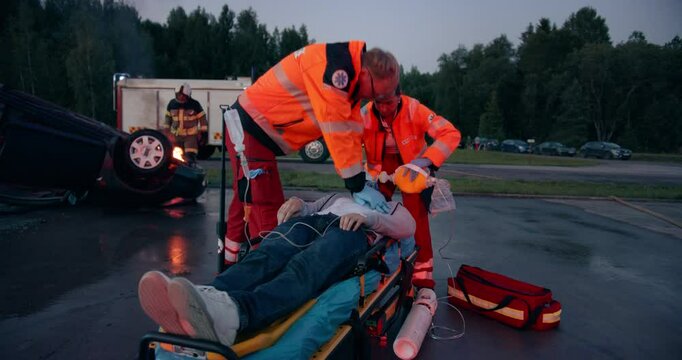 Ambulance Medics Doing CPR to an Unconscious Patient After a Serious Road Accident with a Rolled Car. Scene with Burning Car, Firefighters and Fire Engines in the Background