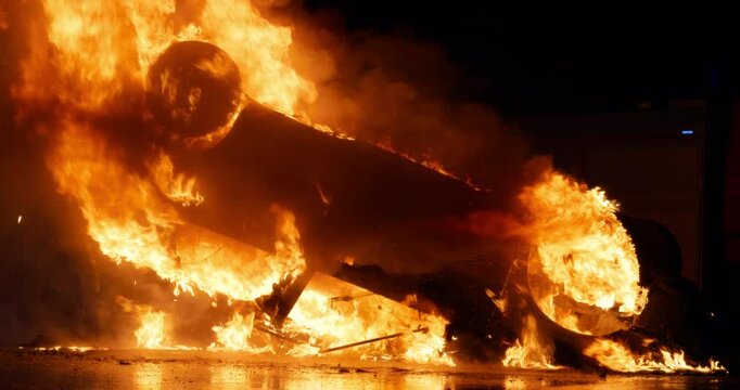 Car Accident with an Overturned Vehicle Engulfed in Flames After a Crash. Emergency Responders Arrive on the Scene, Ready to Battle Intense Fire Late at Night. Footage Without People