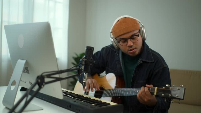 Young asian musician composing a song with playing guitar and piano music instrument in digital recording home studio, a composer man mastering music audio for soundtrack with piano keyboard.
