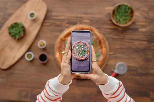 Top view of unrecognizable woman taking photo of fresh delicious pizza and holding smartphone over wooden table during mobile food photography set copy space