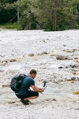 a man in hiking clothes and with a backpack squats by a mountain stream, filming it with a camera mounted on a stabilizer. It is surrounded by rocky shores and clear waters, with trees in the backgrou