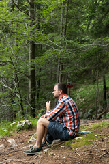 a man squatting on a forest path. He is wearing a plaid shirt and shorts, holding a pipe or electronic device in his hand. It is surrounded by dense green forest, creating a calm and secluded atmosphe