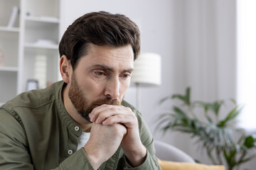 Pensive man in casual clothing sits thoughtfully at home, hands clasped near face. Capturing mood...