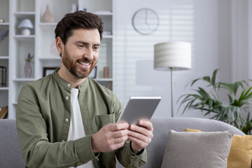Smiling man in casual attire holding tablet while sitting on sofa. Bright and cozy living room setting conveying feelings of relaxation, technology connection, and personal time.