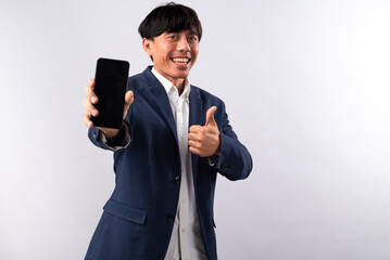 Naklejka premium An Asian man in a navy blue suit confidently while holding a smartphone towards the camera, giving a thumbs-up gesture with his other hand. He stands against a plain white background.