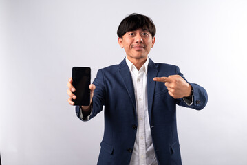 Naklejka premium An Asian man in a blue suit confidently points at a smartphone he is holding, emphasizing the device. He stands against a plain white background, highlighting a professional