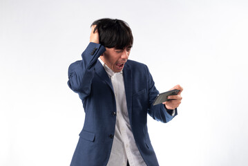 An Asian man in a blue suit expresses shock and frustration while looking at his smartphone, with a hand on his head. He stands against a plain white background, enhancing the emotional intensity