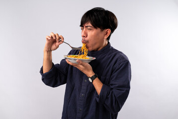 An Asian man in a dark shirt eagerly eats noodles from a plate, his expression filled with delight....