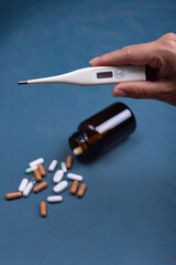 A hand holds a thermometer while some pills are placed on a blue background.