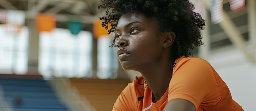 A young Black woman a female basketball player is seen in a side view portrait sitting on a bench in an indoor court with flags in the background providing copy space for an image - Powered by Adobe