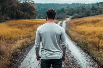 A man wearing a gray sweater stands alone on a winding rural path surrounded by picturesque countryside, with a scenic background of fields and distant trees in autumn colors.