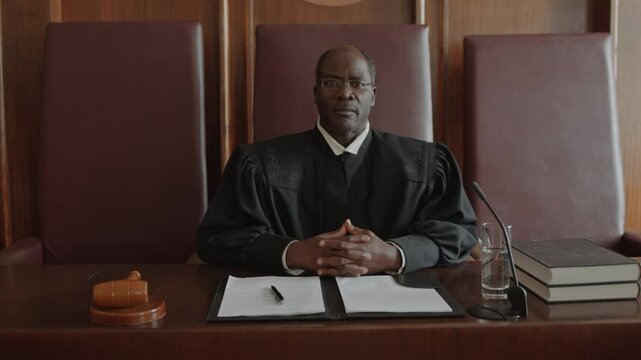 Portrait of experienced African American male judge in eyeglasses and black mantle looking at camera while sitting at judges bench in courtroom