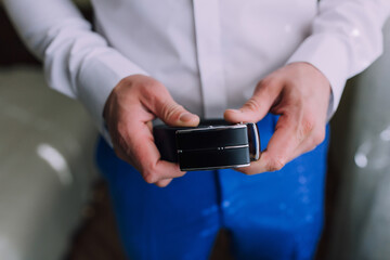 A man is wearing a blue belt and holding it up to his waist. Concept of formality and attention to detail, as the man is dressed in a suit and tie