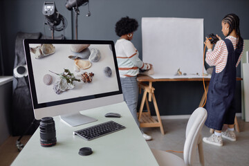PC computer with product photography pictures on screen at workplace in photo studio two girls taking photos in background copy space
