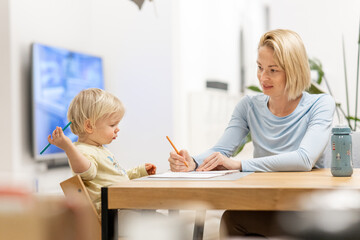 Caring young Caucasian mother and small son drawing painting in notebook at home together. Loving mom or nanny having fun learning and playing with her little 1,5 year old infant baby boy child