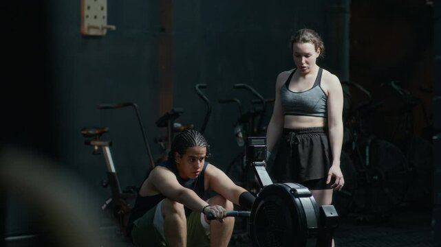 Female fitness instructor watching young man using rowing machine and correcting his technique