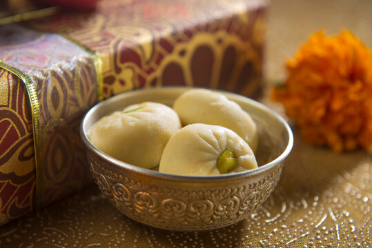 A bowl of an Indian sweet along with festive gift box in the background. Pedha- a traditional Indian sweet made with milk, served in a silverware.