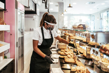 Beautiful young female worker with protective mask  and face shield working in bakery. Business and coronavirus pandemic concept.