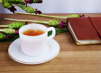 Red-brown hot tea in a ceramic cup and saucer sits between books and flowers on a wide wooden table.

