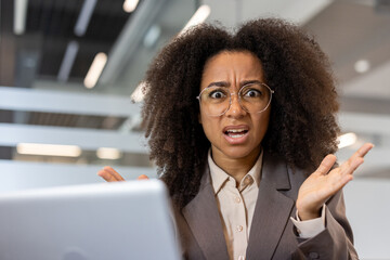 Close-up portrait of a young African-American angry woman sitting in the office at her desk and casually waving her arms at the camera