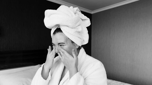 Horizontal 4K black and white video. Young adult white woman in robe and towel on head applies milk balm to face in cozy hotel bed room. 14 february, march 8, spa salon, skin care, rest, relax