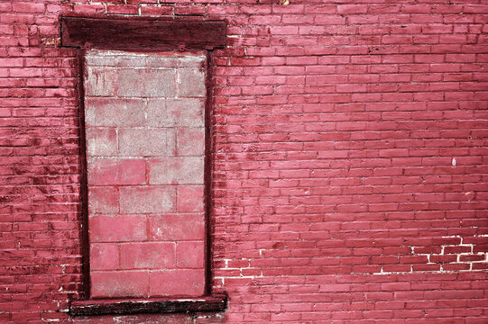 A weathered red brick wall featuring an old window opening that has been completely sealed and bricked up with light colored concrete blocks in an urban building facade.